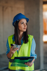 Female engineer using smartphone and holding clipboard on construction site