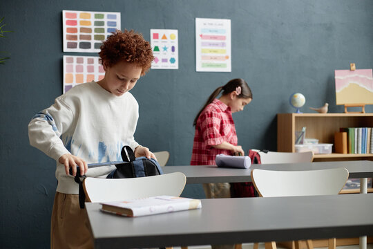 Caucasian boy and Caucasian girl standing in classroom packing backpacks on desks, focusing on organizing school supplies, educational posters and books visible in background - Powered by Adobe
