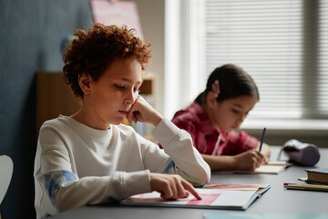 Caucasian boy reading book at desk while teenage girl with dark hair writing in notebook in...