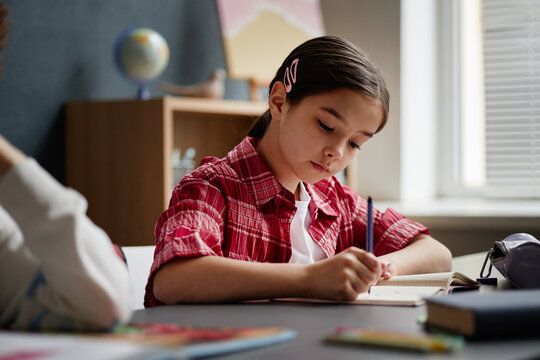 Caucasian girl child sitting at desk writing in notebook with focused expression, long dark hair pulled back with hair clip, classroom setting with globe and books visible in background - Powered by Adobe