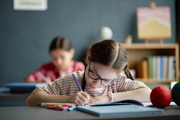 Caucasian girl child wearing glasses sitting at desk writing in notebook with colored pencils in...