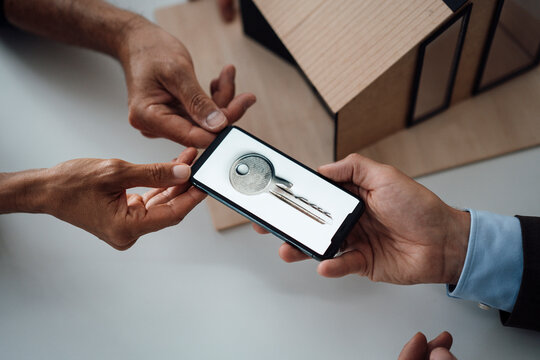 Hand of real estate agent giving mobile phone with house key on screen to customers at table