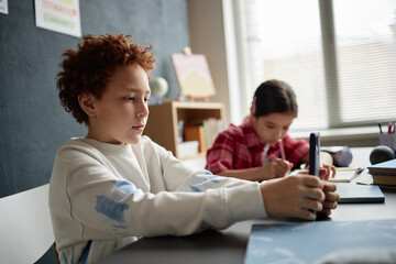 Caucasian boy using smartphone at desk while teenage girl with dark hair writing in notebook in classroom setting, both focused on individual tasks during school activity