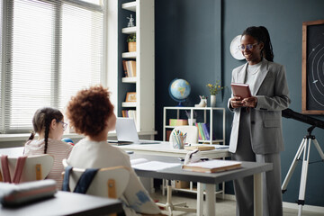 Young adult Black female teacher standing at front of classroom holding notebook, engaging with two seated children, one Caucasian boy and one Caucasian girl, during astronomy lesson