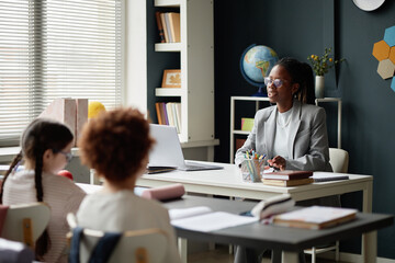 Black female teacher sitting at desk talking to two children in classroom, using laptop while...