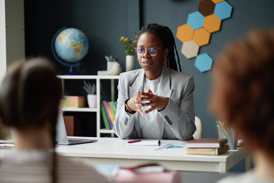 Young Black female teacher sitting at desk talking to chidren during lesson, hands clasped, wearing glasses, globe and geometric wall decor in background, appearing engaged in conversation