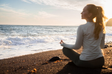 Back side view of a young long haired curly woman looking to the side and meditating on the sea beach at the golden sunset. Girl meditates in yoga asana Padmasana. Female yogi doing yoga outdoors.