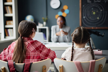 Two Caucasian girls sitting in classroom facing Black female teacher, listening attentively during...