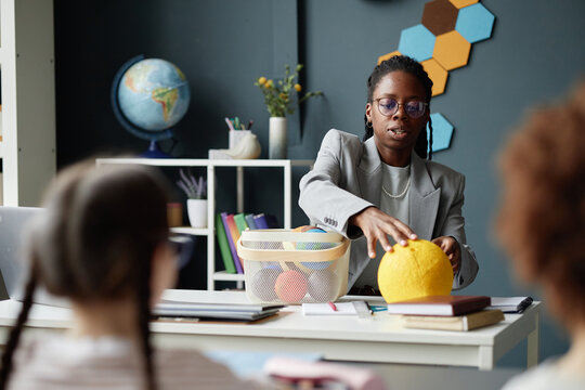Black young adult woman teaching science lesson to group of children in classroom, demonstrating Sun model while students sitting at desk and watching attentively