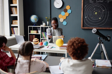Black female teacher demonstrating solar system model to group of diverse children sitting at desks in classroom, children watching attentively, telescope and educational materials visible