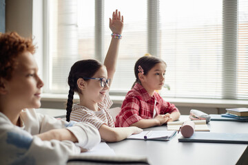 Three children sitting at classroom desk, Caucasian girl with glasses raising hand while smiling,...