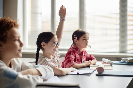 Three children sitting at classroom desk, Caucasian girl with glasses raising hand while smiling, two other children, one Caucasian boy and one Asian girl, listening attentively