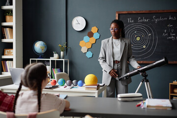 Black woman teacher standing near telescope explaining solar system to children in classroom,...