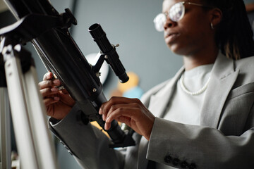 Young adult Black woman adjusting telescope with focused expression, wearing glasses and suit,...