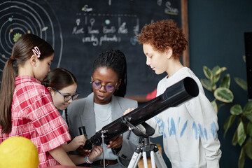 Black woman teaching diverse group of children using telescope in classroom, three girls and one...