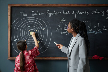 Black woman teacher explaining solar system diagram on chalkboard while Caucasian girl student...
