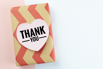 A rectangular gift box wrapped in cream-colored paper with a repetitive red chevron pattern, featuring a pink heart-shaped label with "THANK YOU" on background of pastel pink and white. Top view.