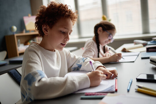 Caucasian boy and Caucasian girl sitting at desk writing in notebooks during classroom lesson, focused on learning, school supplies scattered on table, sunlight streaming through window blinds - Powered by Adobe