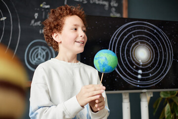Caucasian boy holding model of Earth while standing in front of solar system diagram on screen,...