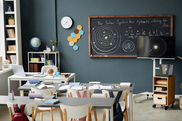 Empty classroom showing desks with school supplies, backpacks on chairs, chalkboard and monitor...