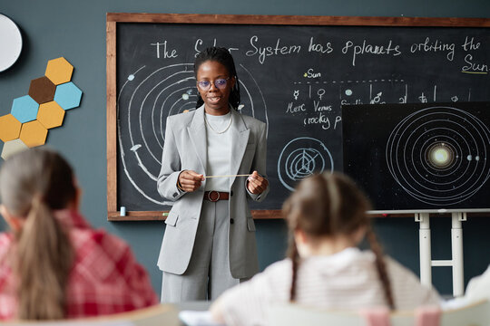 Black woman teacher explaining solar system diagram to group of children in classroom, holding pointer and standing in front of chalkboard with planetary drawings, students listening attentively
