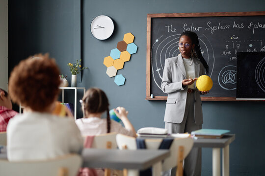 Black woman teacher explaining solar system model to group of children in classroom, holding model of Sun while students sitting at desks listening and looking toward chalkboard