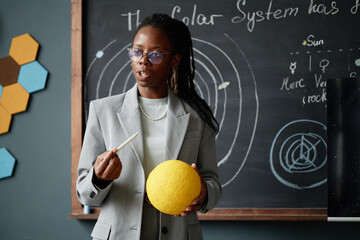 Young adult Black woman standing in classroom holding yellow model of Sun and pointing with chalk,...