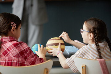 Two Caucasian girls sitting at table examining textured model planets with hands, engaging in...