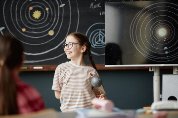 Caucasian girl child with glasses standing in classroom holding model planet, smiling and looking...