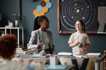Black woman teacher interacting with Caucasian girl student holding model planet in science...
