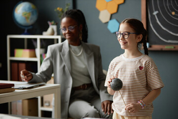 Caucasian girl smiling and holding model planet while standing in classroom, Black woman teacher...