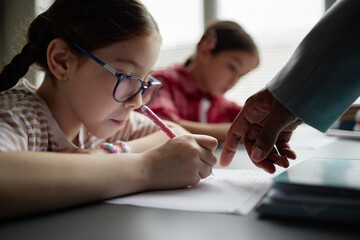 Caucasian girl child wearing glasses writing in notebook while hand of Black female teacher...