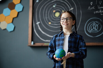Caucasian girl child with brown hair and glasses standing in classroom holding model of Earth,...
