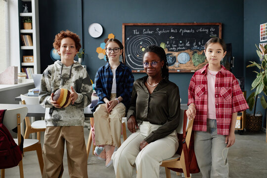 Caucasian boy holding planet model standing beside Caucasian classmate, Black female teacher sitting on chair, Asian girl standing in astronomy classroom with chalkboard showing solar system diagram - Powered by Adobe