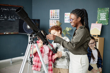 Black young adult woman demonstrating telescope to group of diverse children in classroom, children...