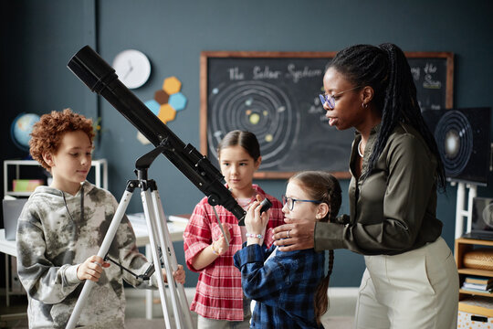 Black woman teaching group of children using telescope in classroom, three kids including Caucasian boy and two girls of different ethnicities observing astronomy lesson together