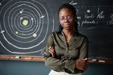 Portrait of young adult Black woman standing with arms crossed in front of chalkboard showing solar...