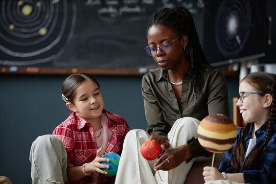 Black woman teaching two preteen girls about solar system models, holding planet replicas while children listening and interacting, classroom setting with chalkboard in background