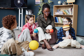 Black woman teaching diverse group of children about planets, sitting on floor in classroom, children holding model planets, engaging in interactive science lesson together