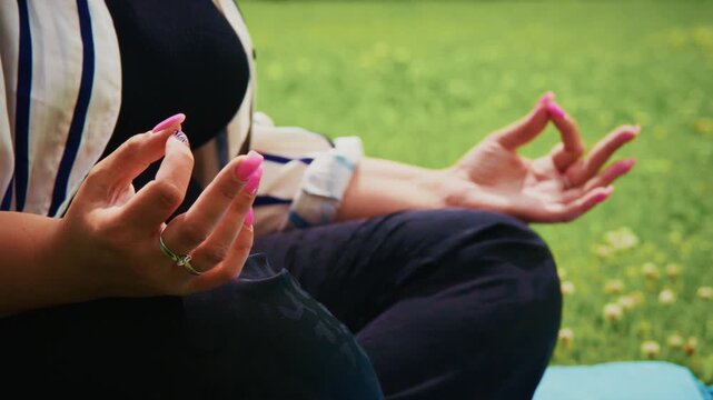 Woman engaged in meditation outdoors, sitting cross-legged on a yoga mat, with hands in mudra position, surrounded by lush green grass and a serene natural environment, promoting mindfulness
