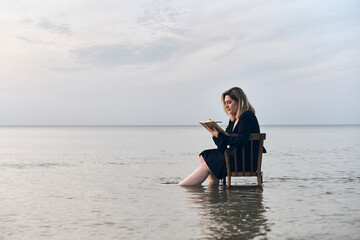 Young woman sits on chair in shallow water at sunset, writing in notebook. Serene ocean setting...