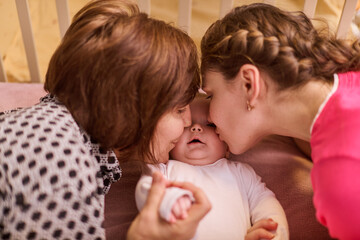 Elderly woman with brown hair and young woman with braided hair, both affectionate, lean toward infant inside crib. Soft lighting, warm, intimate atmosphere
