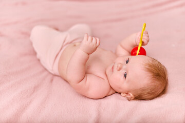 Baby girl with light brown hair and fair skin plays on soft pink blanket. Gaze focused on red toy,...