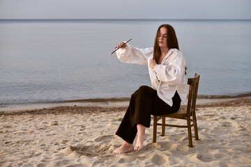 Young female artist enjoys quiet reflection on beach, holding paintbrush. She sits barefoot in...