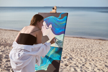 Young female artist paints vibrant portrait by seaside, capturing serene ambiance of beach. Her...