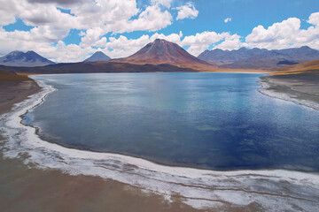The stunning landscape features a serene lake surrounded by mountains, under a blue sky adorned with fluffy clouds, set against the backdrop of a vast desert