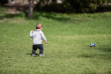 Fototapeta premium Baby walking on grass towards ball in park