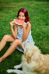 Teen girl enjoying watermelon while sitting on grass with a dog nearby during a sunny day in the backyard
