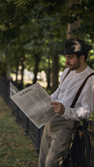 Man in vintage attire reads newspaper while leaning against a park fence under trees. Concept of...