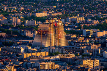Panoramic view of the Parisian cityscape with a modern skyscraper under construction, tall cranes, and the golden light of sunset illuminating everything.